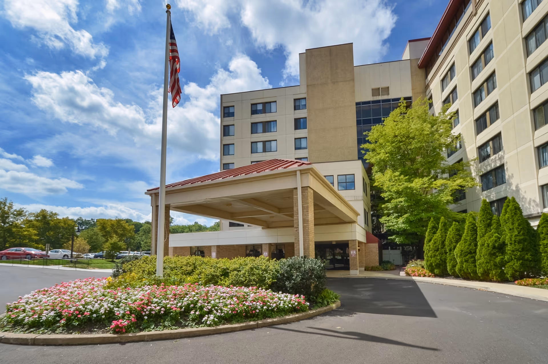 Exterior view of Wesley Enhanced Living Doylestown building with a covered entrance, an American flag on a flagpole, landscaped flower beds, and trees under a partly cloudy sky.