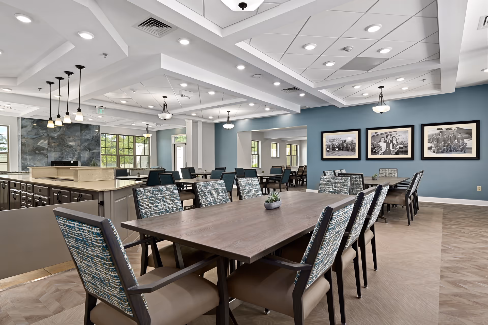 A spacious dining room with multiple wooden tables and cushioned chairs featuring patterned upholstery. The room has a modern design with a blue accent wall adorned with three black-and-white framed photographs. Pendant lights hang from the ceiling, and large windows allow natural light to fill the space. There is a kitchen or serving area with a countertop and cabinetry visible on the left side.