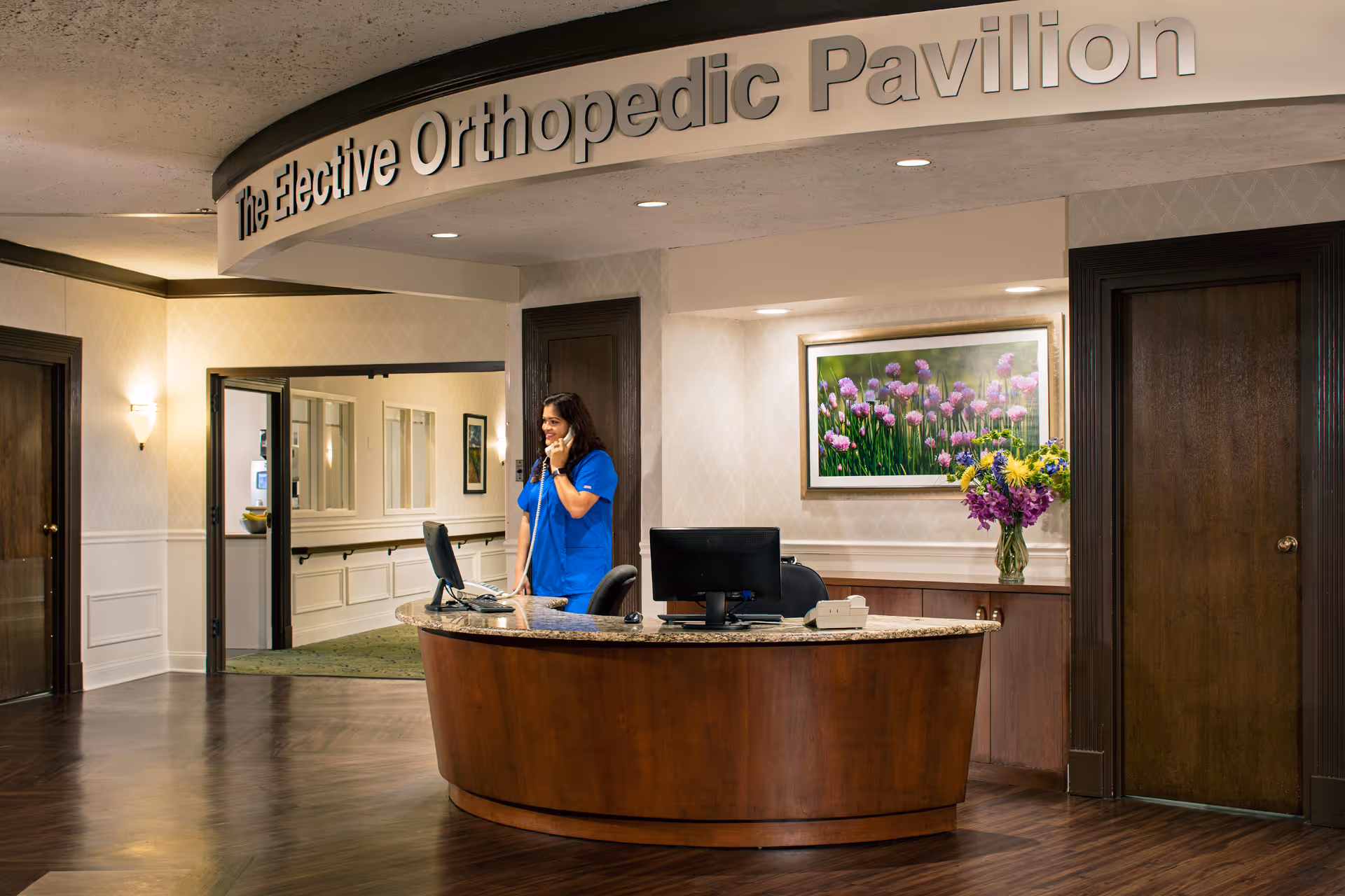 Reception area of The Elective Orthopedic Pavilion with a woman in blue scrubs talking on the phone behind a curved wooden desk with a computer. The area has wooden doors, a framed picture of purple flowers on the wall, and a vase with colorful flowers on a counter behind the desk.