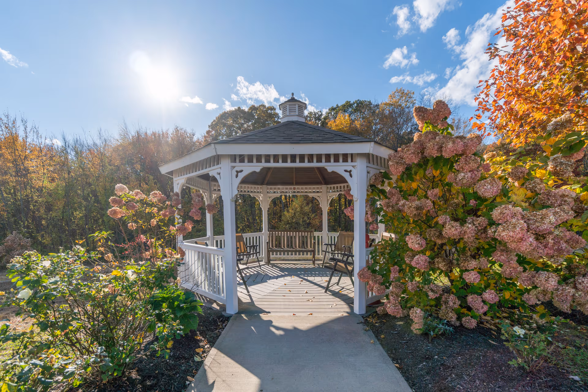 A white wooden gazebo surrounded by flowering bushes and trees with autumn foliage under a bright blue sky with scattered clouds and sunlight.