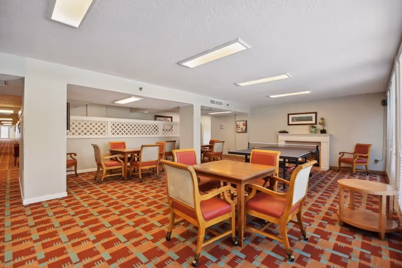 A common area in a senior living facility featuring several wooden tables with cushioned chairs arranged around them. The room has a patterned carpet in shades of red, orange, and beige. There is a ping pong table near the back wall, which also has a white mantel with decorative items and framed artwork above it. Large windows with vertical blinds allow natural light to enter the room.