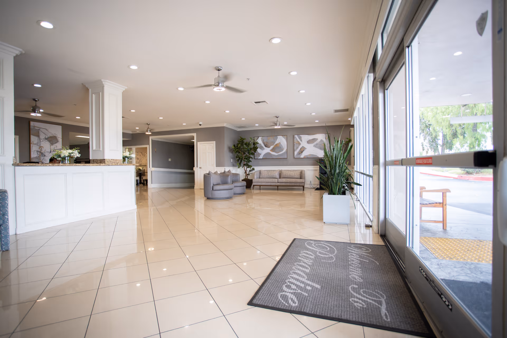 Bright and spacious lobby area of a senior living facility with tiled floors, a white reception desk on the left, modern seating including a sofa and two armchairs, large potted plants, abstract artwork on the walls, ceiling fans, and large glass doors leading outside with a welcome mat that reads 'Welcome to Paradise'.