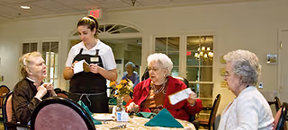 Three elderly women seated around a dining table in a retirement community, engaged in conversation, while a waitress stands beside them taking an order. The table is set with napkins, plates, and a small flower arrangement. The room has large windows and a warm, inviting atmosphere.