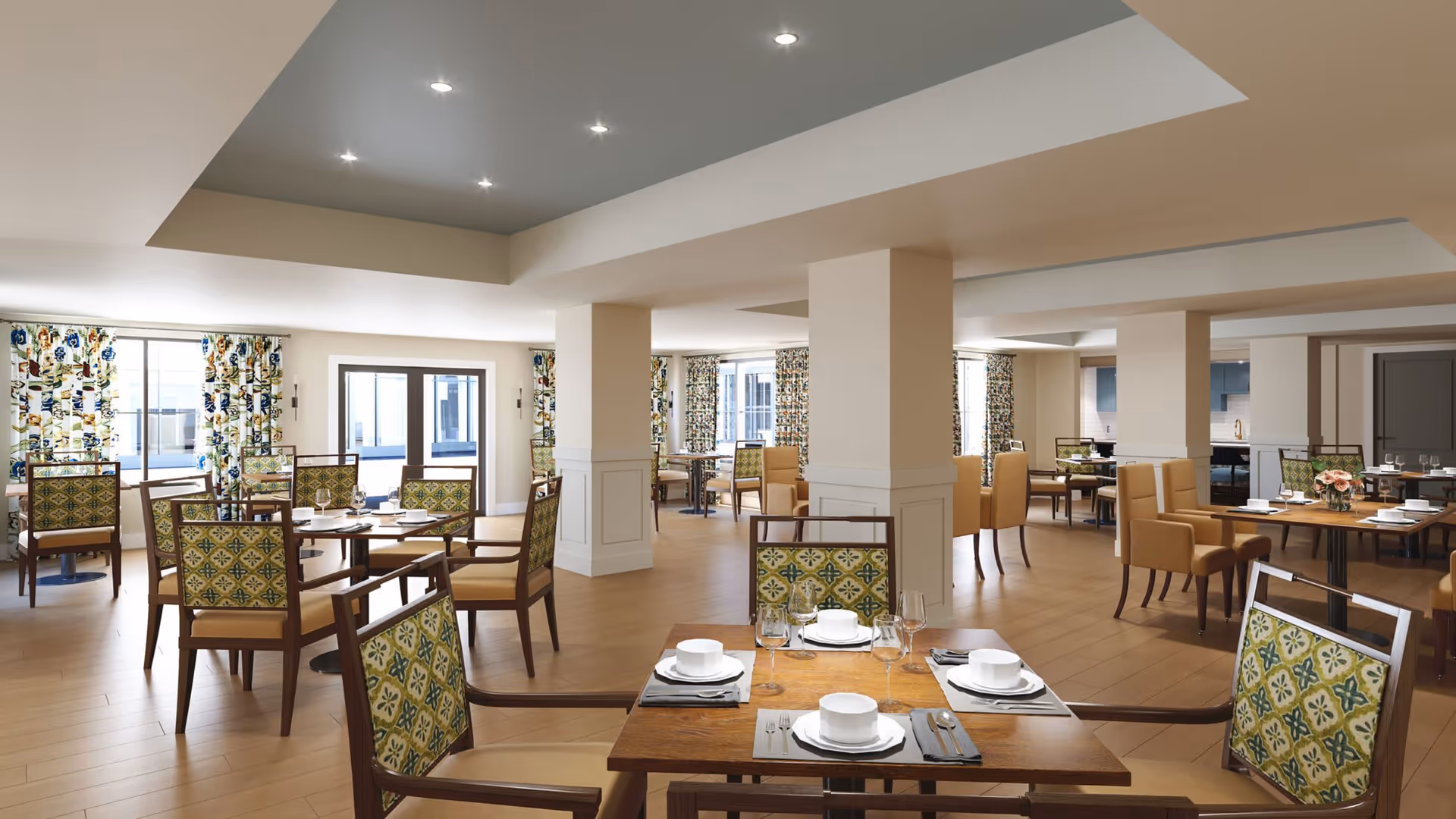 A spacious dining room with wooden tables set with white plates, bowls, glasses, and silverware. The chairs have patterned green and yellow upholstery, and some have solid tan upholstery. The room has large windows with floral curtains, light wood flooring, and recessed ceiling lights.