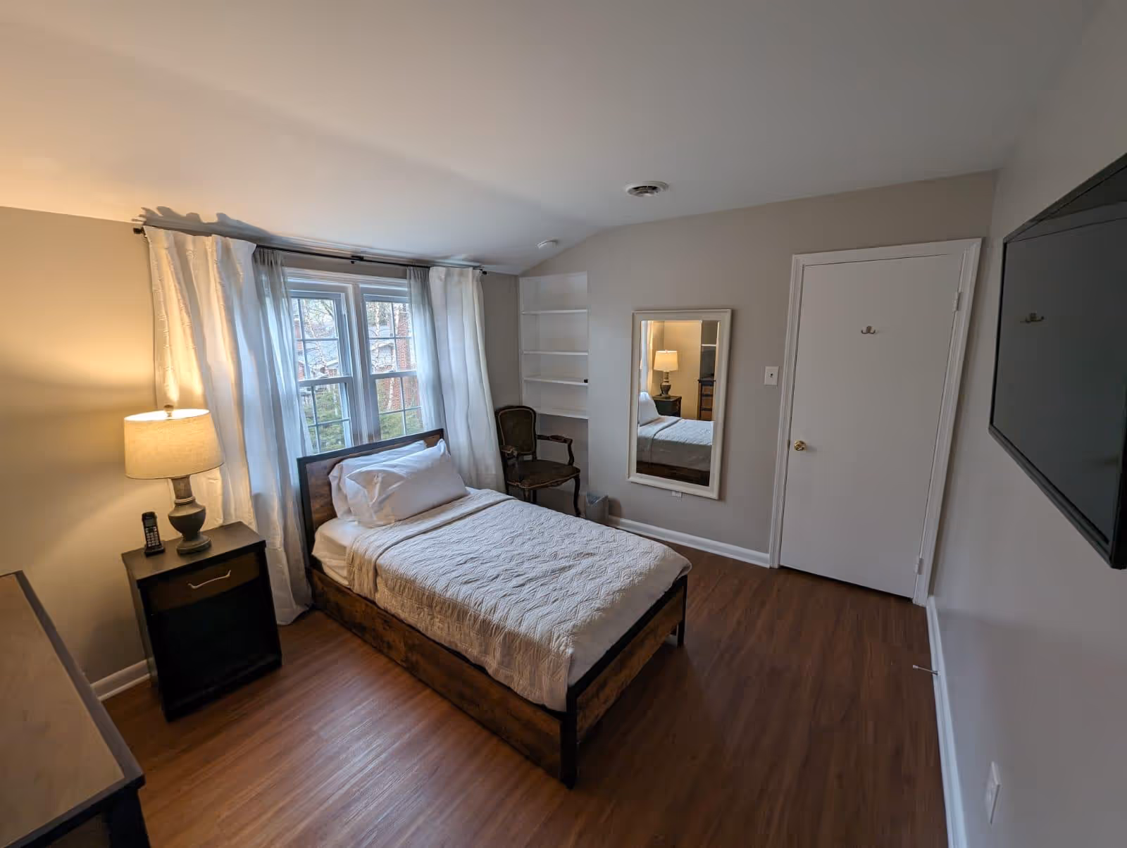 A small, neatly furnished bedroom with a wooden bed, nightstand, lamp, window, and mirror.
