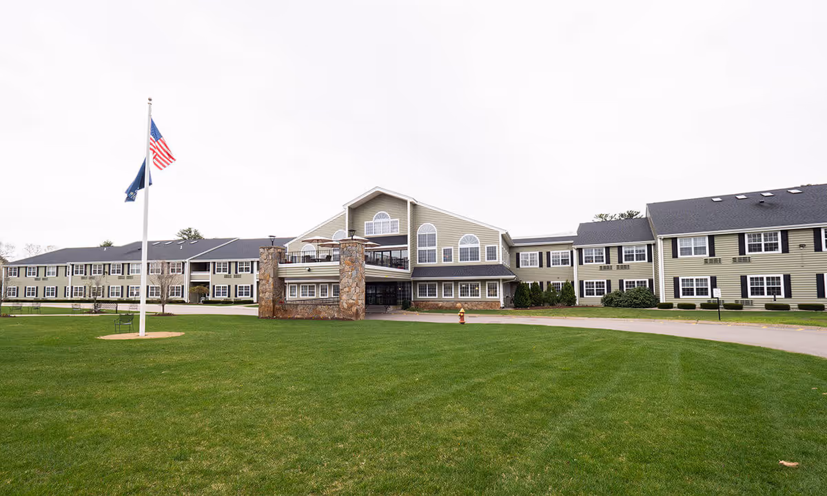 Exterior view of a large senior living facility building with beige siding, black shutters, and a dark roof. The building has multiple windows and a central entrance area with stone pillars and a covered porch. In front of the building is a well-maintained green lawn with two flagpoles flying the American flag and another flag.