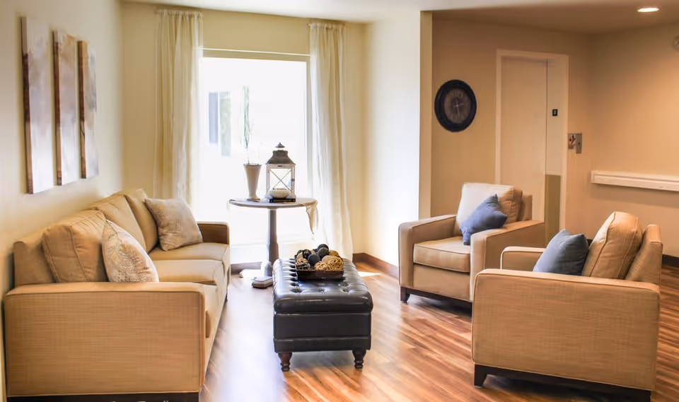 A cozy senior living facility common area with a beige sofa, two beige armchairs with blue cushions, a black tufted ottoman with decorative items on top, a round side table with a lantern and vase, wooden flooring, and a large window with sheer curtains letting in natural light.