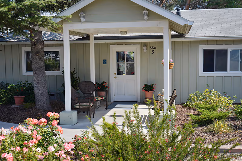 Covered front entrance of a single-story building with a white door, porch chairs, potted plants, and flowering shrubs.