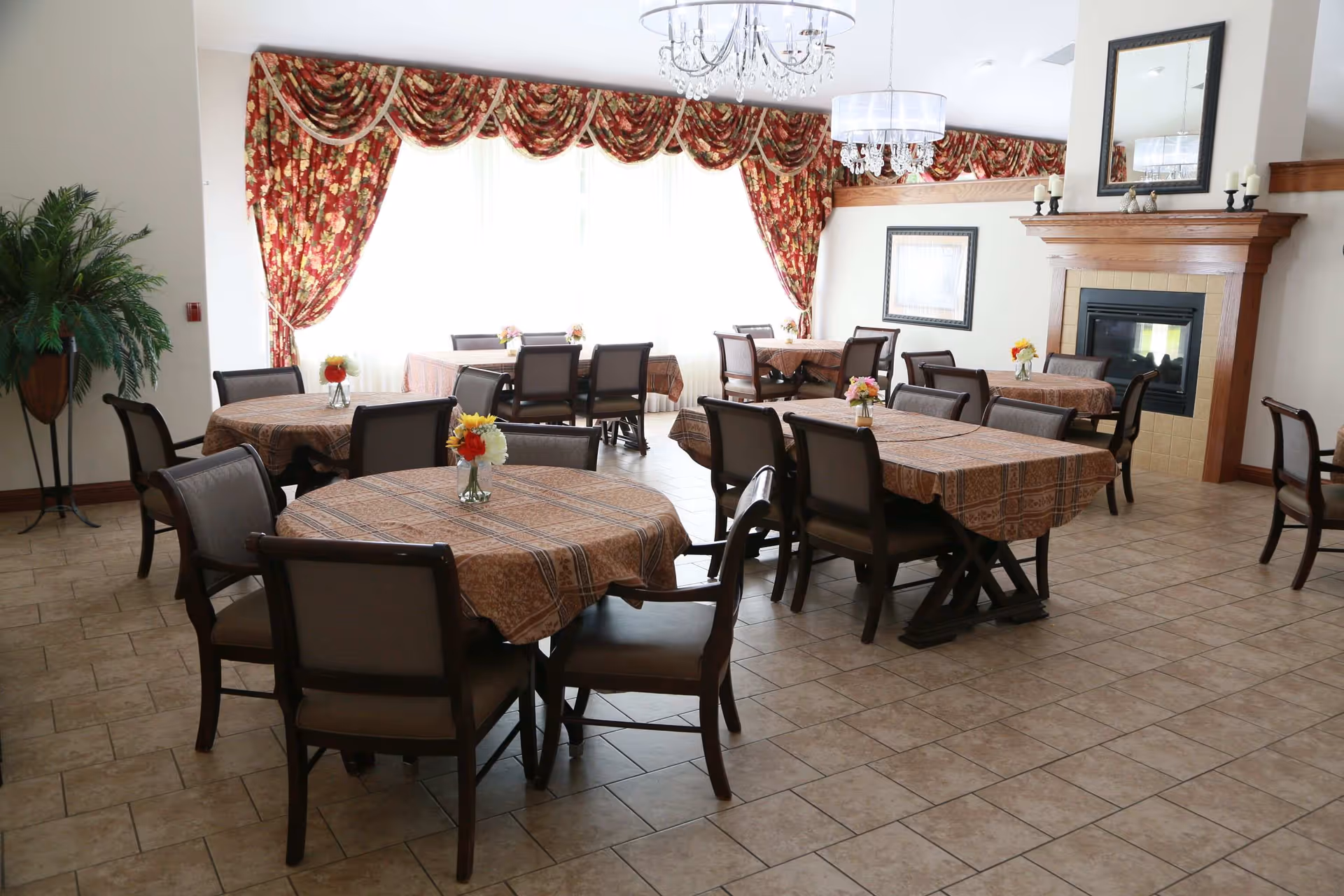 A dining room with several tables covered in patterned tablecloths, each with a small vase of flowers. The room has tiled floors, a fireplace with a wooden mantle and mirror above it, floral curtains on large windows, and two chandeliers hanging from the ceiling.