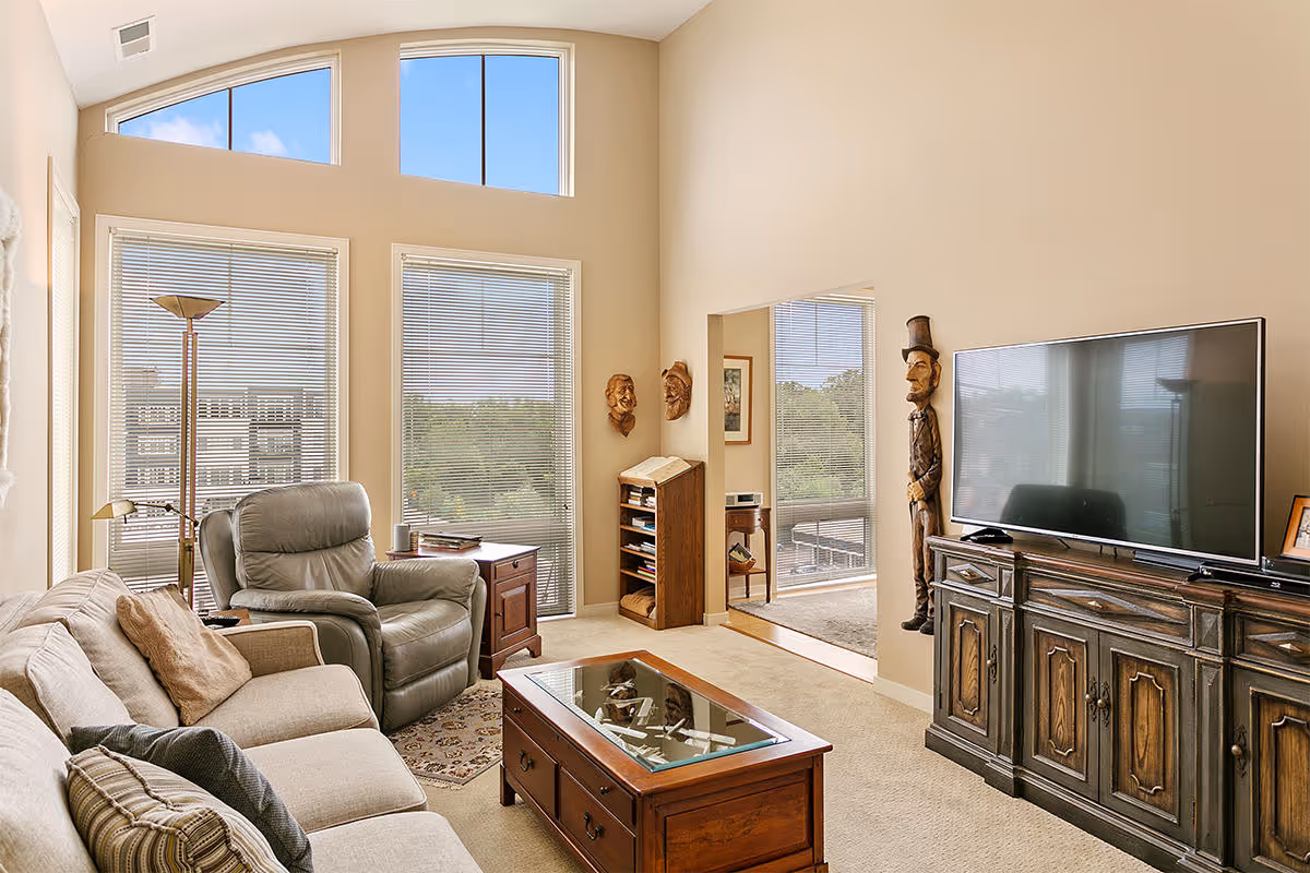 A cozy living room with a beige sofa and a gray recliner chair facing a large flat-screen TV on a wooden cabinet. The room has tall windows with blinds, letting in natural light, and a glass-top wooden coffee table in the center. There are decorative wooden masks on the walls and a wooden sculpture of a man wearing a top hat next to the TV cabinet.