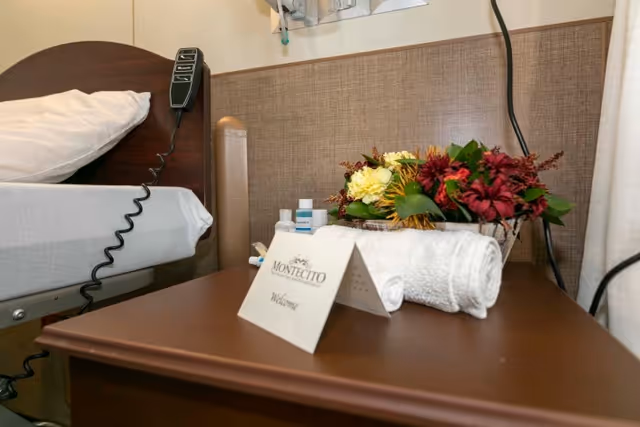 Bedside table in a patient room holding a Montecito welcome card, rolled towel, toiletries, and a floral arrangement beside a hospital bed.