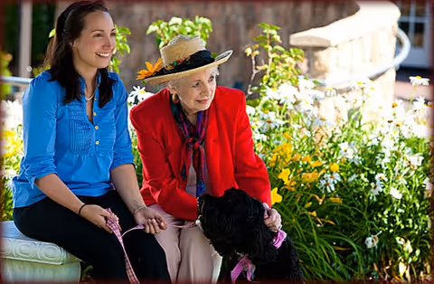 A young woman and an elderly woman sitting outdoors on a bench surrounded by greenery and flowers. The elderly woman is wearing a red jacket and a hat with a flower decoration, and they are both holding a black dog on a leash.