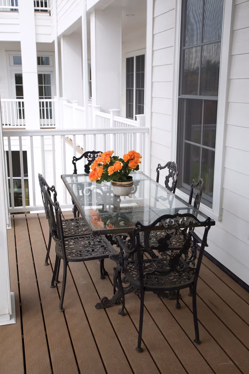 Outdoor balcony area with a glass-top table and six ornate black metal chairs. A potted plant with orange flowers is placed in the center of the table. The balcony has white railings and is attached to a white building with windows.