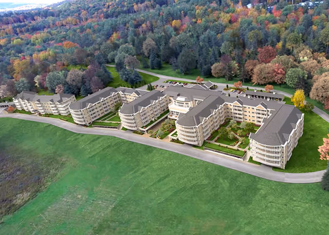 Aerial view of a large senior living facility complex surrounded by green lawns and dense trees with autumn foliage.