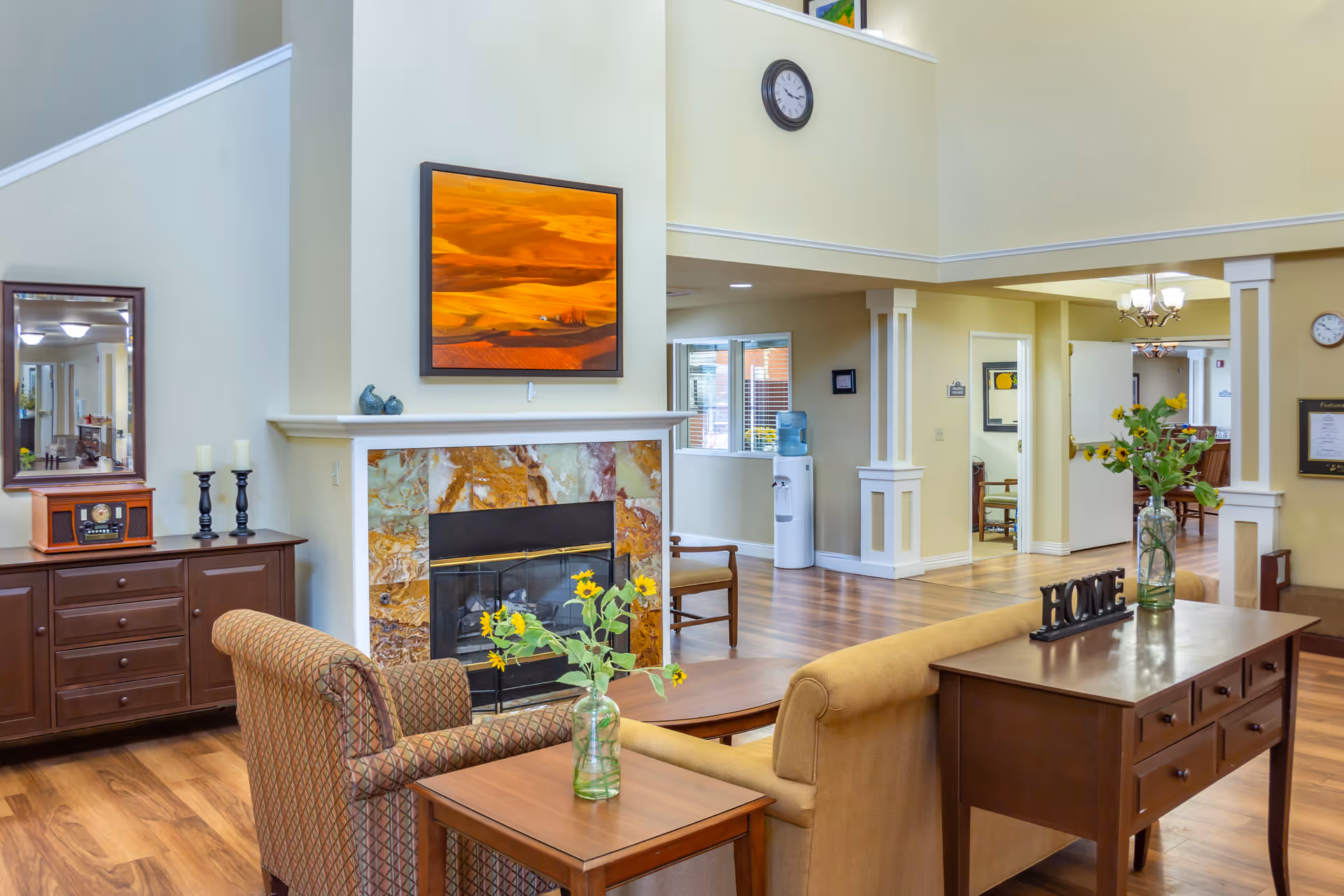 A cozy living room area in a senior living facility with a fireplace featuring a marble surround, a framed landscape painting above it, and a clock on the wall. The room has wooden flooring, a patterned armchair, a beige sofa, and wooden tables with vases of sunflowers. There is a wooden sideboard with candles and a vintage-style radio, and a water cooler is visible in the background near a hallway.