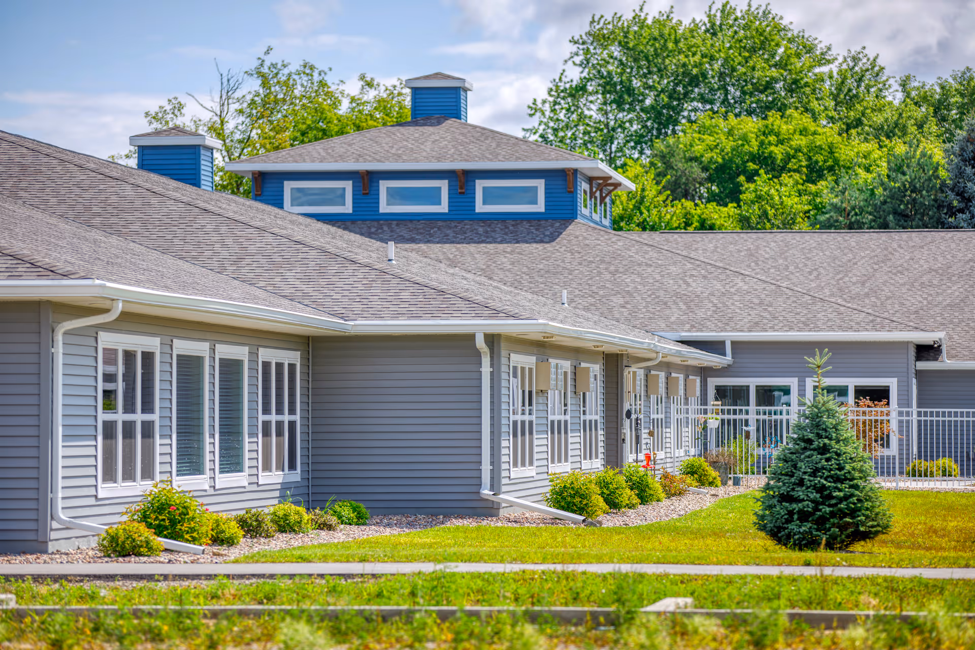 Exterior view of a single-story senior living facility building with gray siding, multiple windows, and a landscaped lawn with bushes and a small evergreen tree under a blue sky.