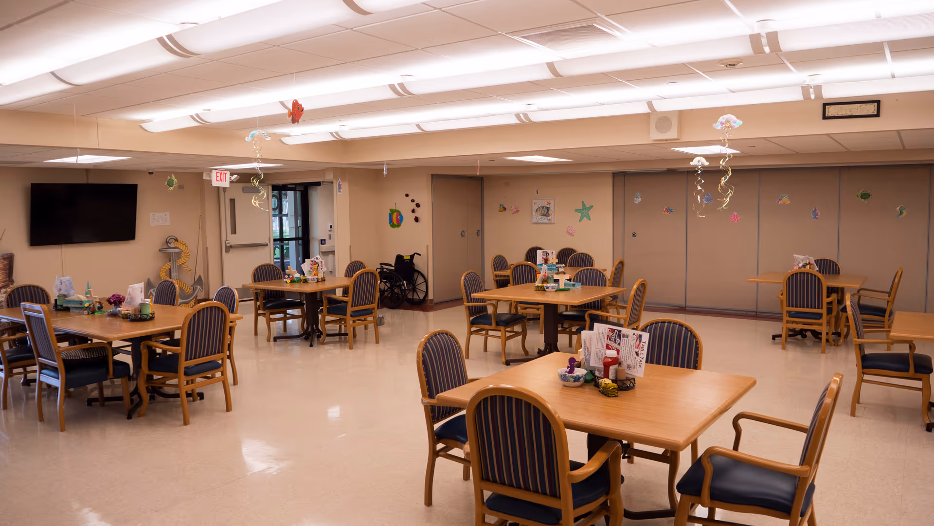 A spacious dining room with several wooden tables and chairs arranged neatly. The chairs have blue and white striped cushions. The room has a polished floor and a ceiling with bright fluorescent lights. Decorations hang from the ceiling and colorful wall decorations are visible on the far wall. A wheelchair is parked near the back wall, and a large flat-screen TV is mounted on the left wall.