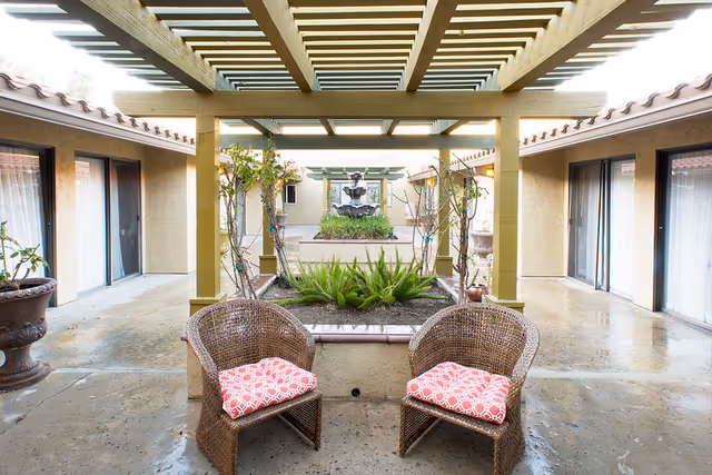 Outdoor courtyard area with two wicker chairs featuring red patterned cushions under a pergola. The courtyard has potted plants, a central garden bed with green plants, and a multi-tiered water fountain in the background. Surrounding the courtyard are beige walls with sliding glass doors.