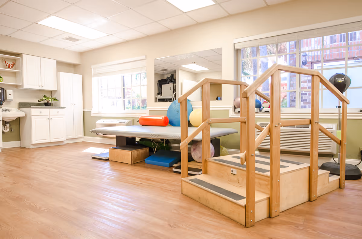 Bright therapy room with wooden practice stairs, a treatment table, exercise balls, and cabinets under large windows.