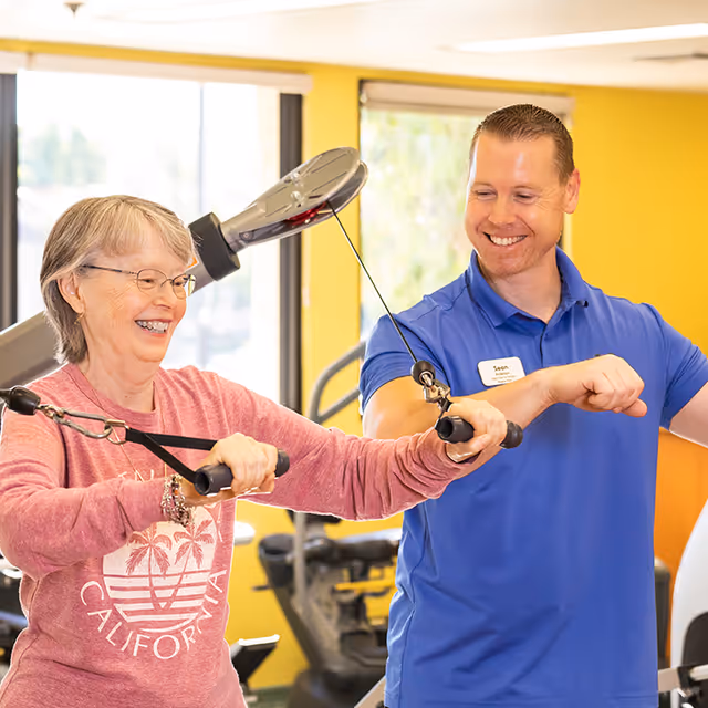 An elderly woman wearing glasses and a pink California sweatshirt is exercising with resistance bands in a gym. A smiling male trainer in a blue polo shirt with a name tag is assisting her. The background shows gym equipment and large windows with natural light.