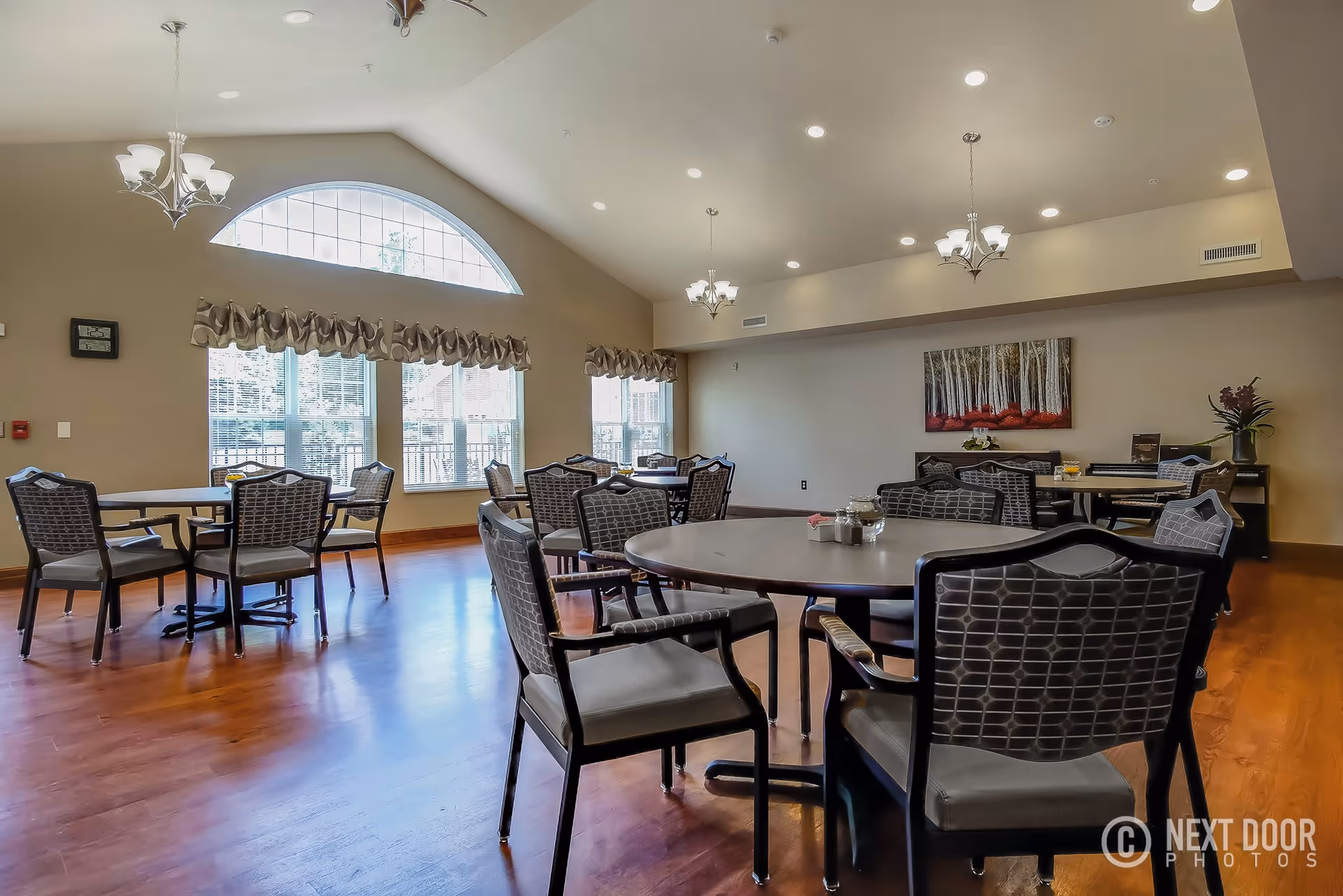 Spacious dining room with multiple round tables and cushioned chairs arranged neatly on a wooden floor. Large windows with decorative valances allow natural light to fill the room, and chandeliers hang from the high ceiling. A painting and some plants decorate the far wall.