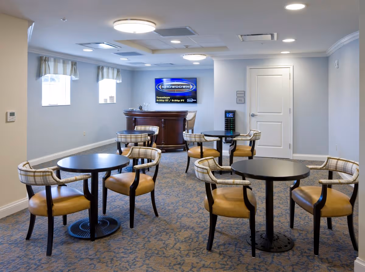 A well-lit common area with four round tables, each surrounded by four cushioned chairs with plaid backs and tan seats. The room has light gray walls, two small windows with striped valances, a wall-mounted TV displaying a show called 'Showdown,' and a wooden bar counter in the back corner. The floor is carpeted with a patterned design.