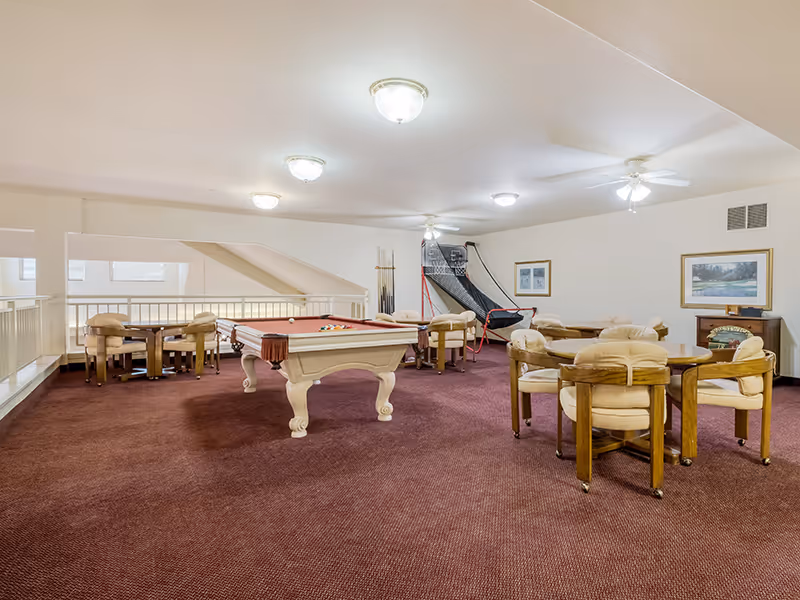 A spacious carpeted recreation room with a pool table, arcade basketball game, and several card tables with cushioned chairs.