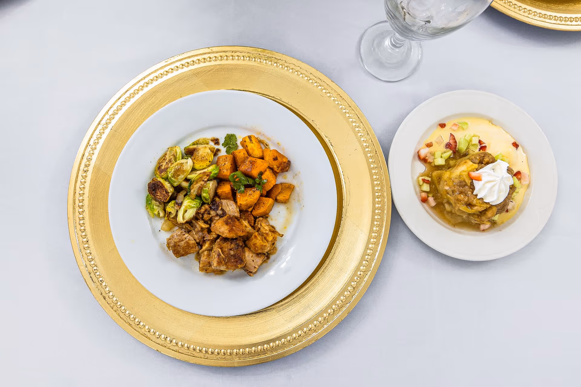 A plated meal on a white tablecloth featuring roasted Brussels sprouts, cubed sweet potatoes, and pieces of cooked meat on a white plate with a decorative gold charger underneath. Next to it is a dessert plate with a pastry topped with whipped cream and surrounded by a creamy sauce with small diced fruit pieces. A clear water glass is partially visible above the plates.