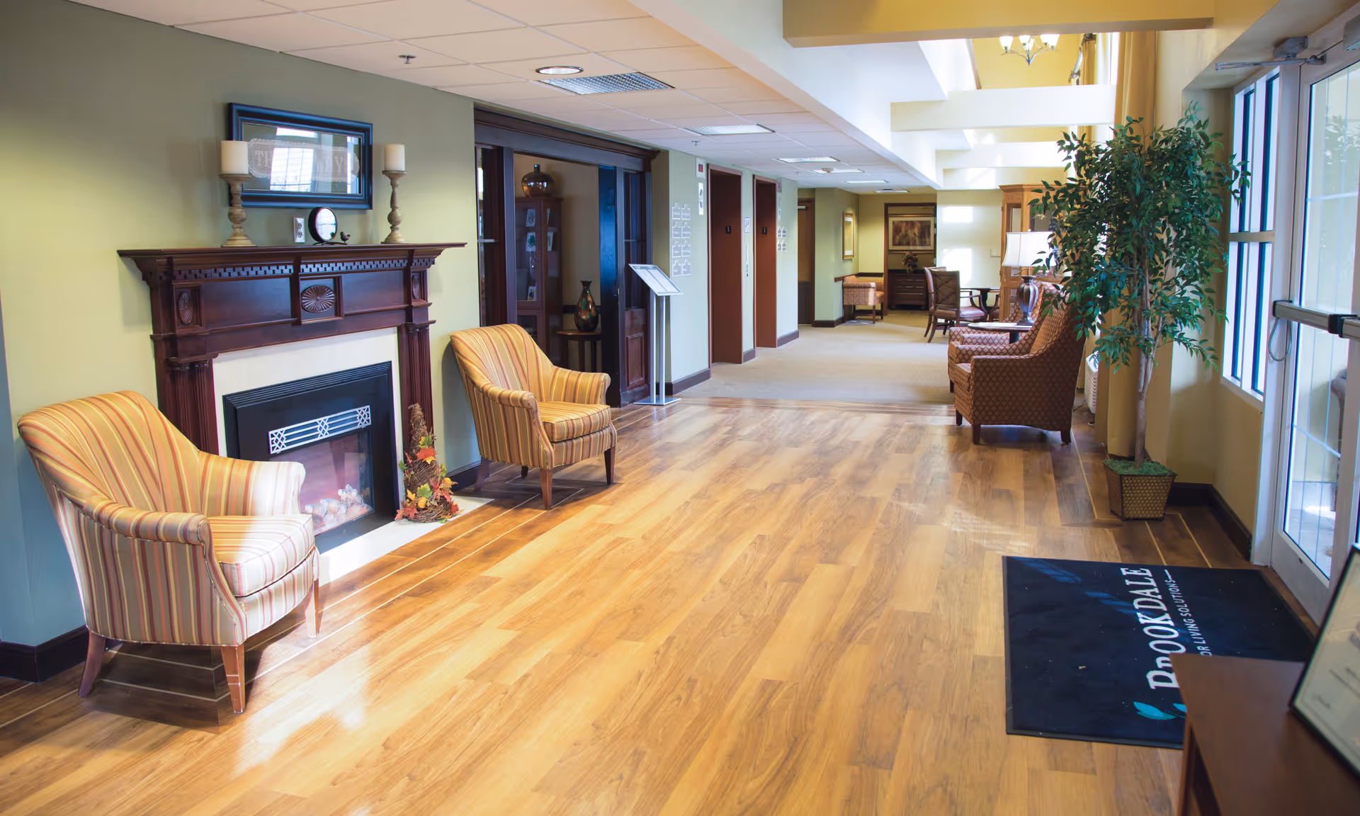 A bright and spacious senior living facility hallway with wooden flooring, two striped armchairs placed near a decorative fireplace with candles and a framed picture above it. The hallway leads to an area with more seating and tables, and large windows on the right side let in natural light. A potted plant is near the entrance door, and a black floor mat with the text 'Brookdale' is visible.