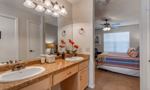 A bathroom with a double sink vanity featuring a granite countertop, two mirrors, and overhead lighting. Decorative items including a vase with orange flowers and soap dispensers are placed on the counter. The bathroom opens into a bedroom with a bed covered in a colorful striped blanket, a ceiling fan with lights, and a window with blinds.