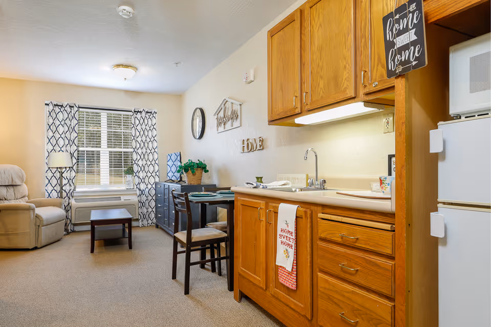 A cozy assisted living apartment interior featuring a small kitchen area with wooden cabinets, a sink, and a white refrigerator with a microwave on top. Adjacent to the kitchen is a dining table set for two with chairs. The living area includes a beige recliner chair, a small coffee table, and a window with patterned curtains letting in natural light. Wall decorations include a clock and signs that say 'HOME' and 'home sweet home'.