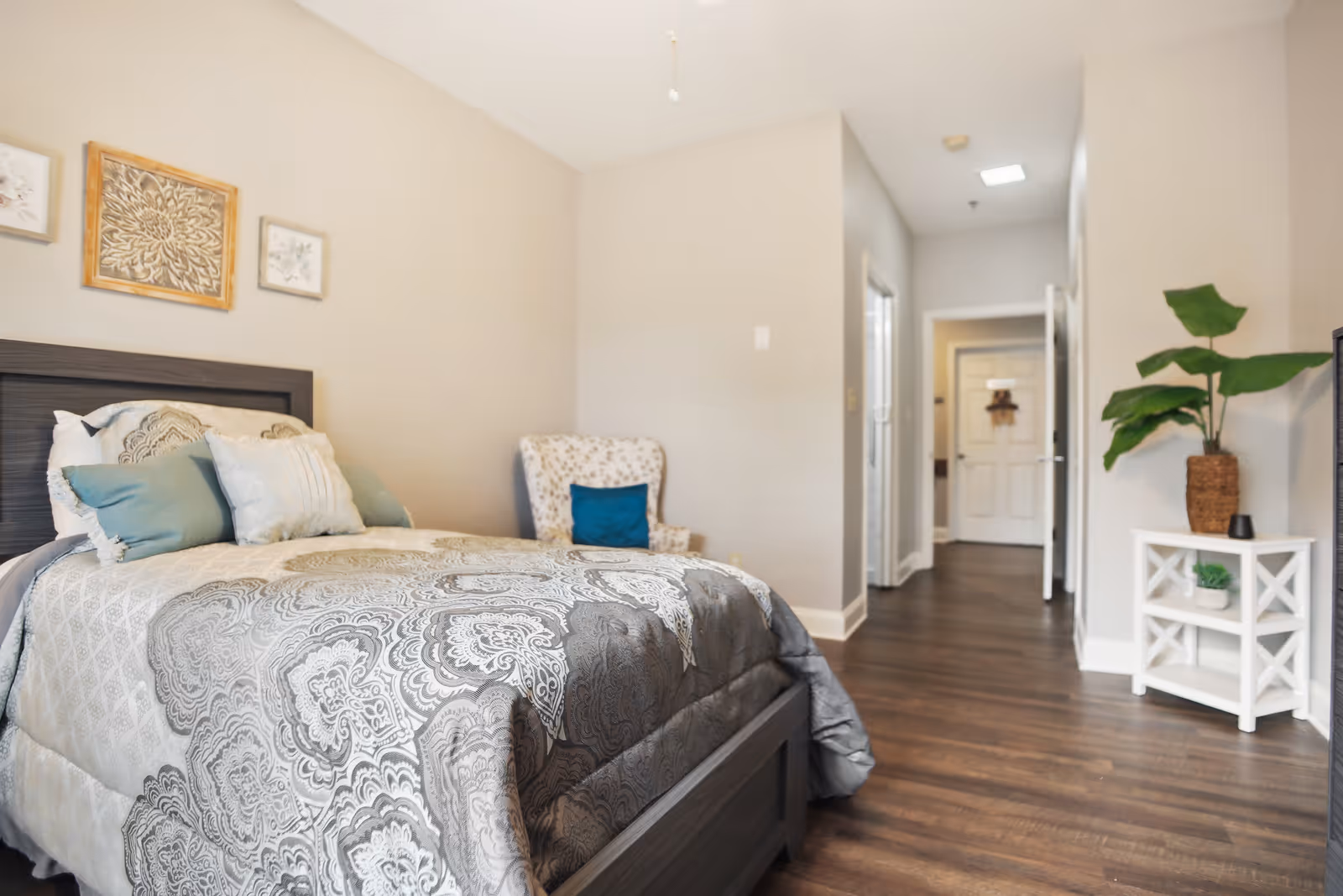 A bright furnished bedroom with a bed, patterned comforter, an accent chair, and a small side table with a plant opening to a hallway.