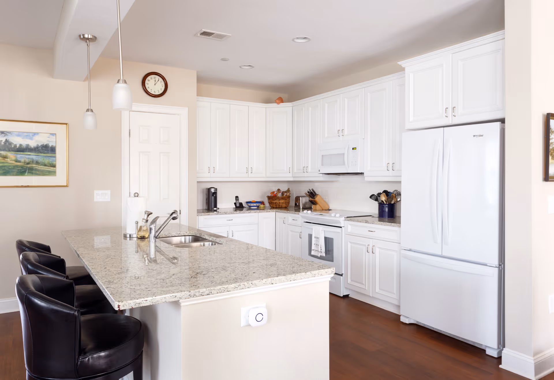 Bright and modern kitchen with white cabinets, a granite countertop island with a sink, three black bar stools, a white refrigerator, a white stove with a microwave above it, and various kitchen utensils and appliances. A clock and a framed landscape painting are visible on the walls.
