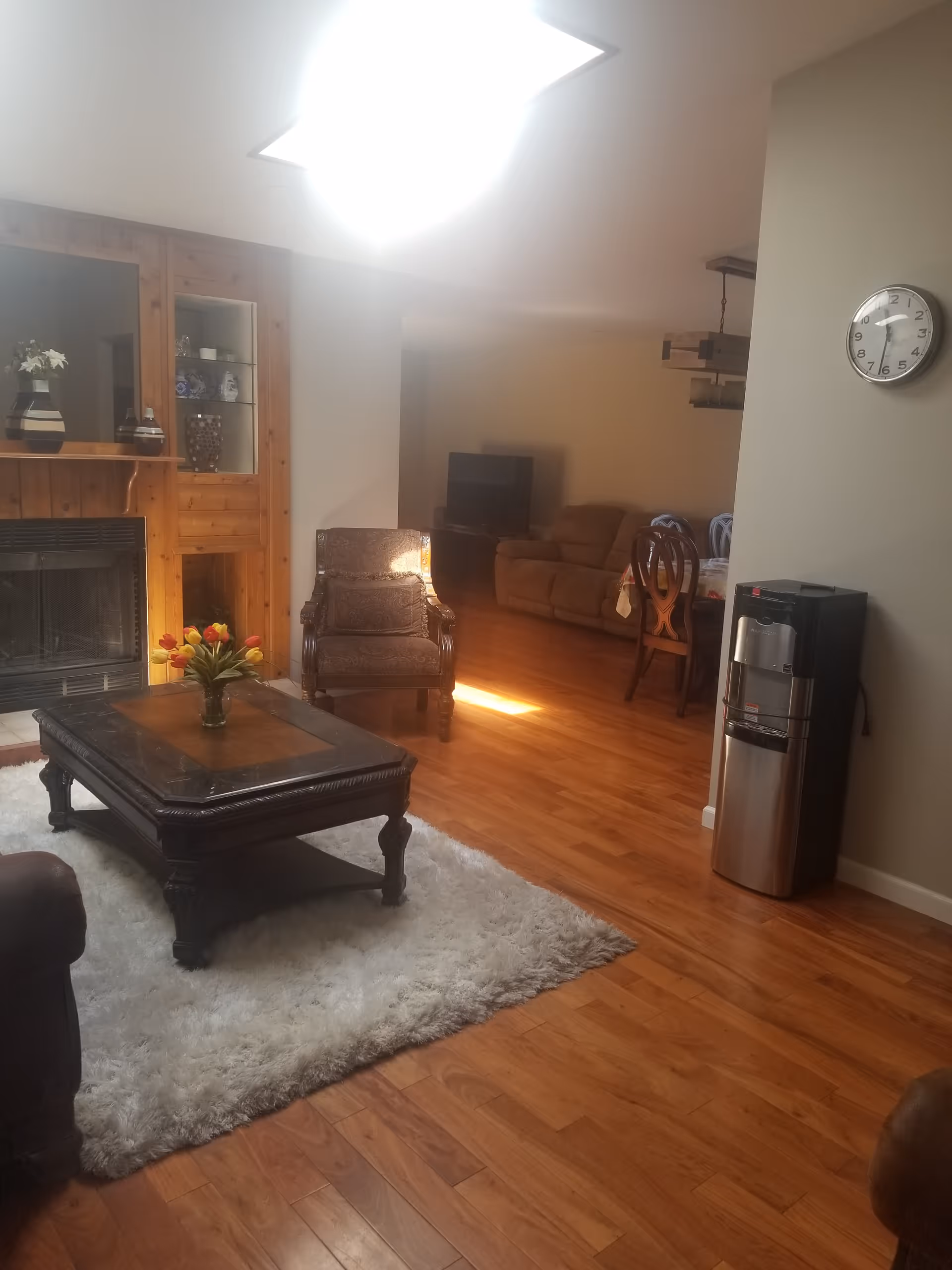 Cozy living room with hardwood floors, a dark wood coffee table on a shag rug, armchairs by a fireplace, and a water cooler and wall clock with a dining area visible in the background.