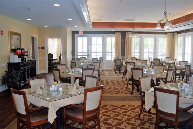 A bright dining room with multiple tables covered with beige tablecloths and set with glasses, napkins, and condiments. The room has large windows with cream-colored curtains, a patterned carpet, and a ceiling with recessed lighting and a chandelier. There is a dark wooden sideboard with coffee dispensers and a plant in the corner.