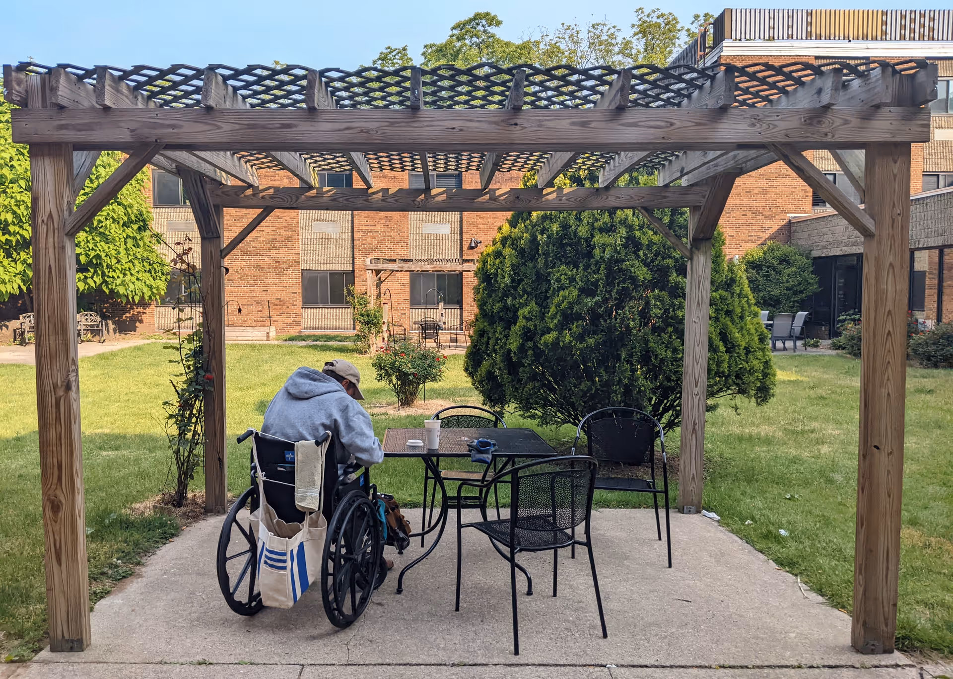 An elderly person in a wheelchair sitting under a wooden pergola in a garden courtyard of a senior living facility. The person is wearing a gray hoodie and a beige cap, facing away from the camera. There is a black metal table with three chairs around it, and greenery including bushes and trees surround the area. The brick building of the facility is visible in the background.
