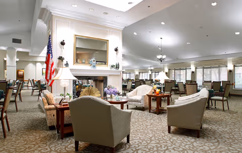 Spacious, well-lit communal dining and lounge area with sofas, armchairs, dining tables, a central fireplace, and large windows.