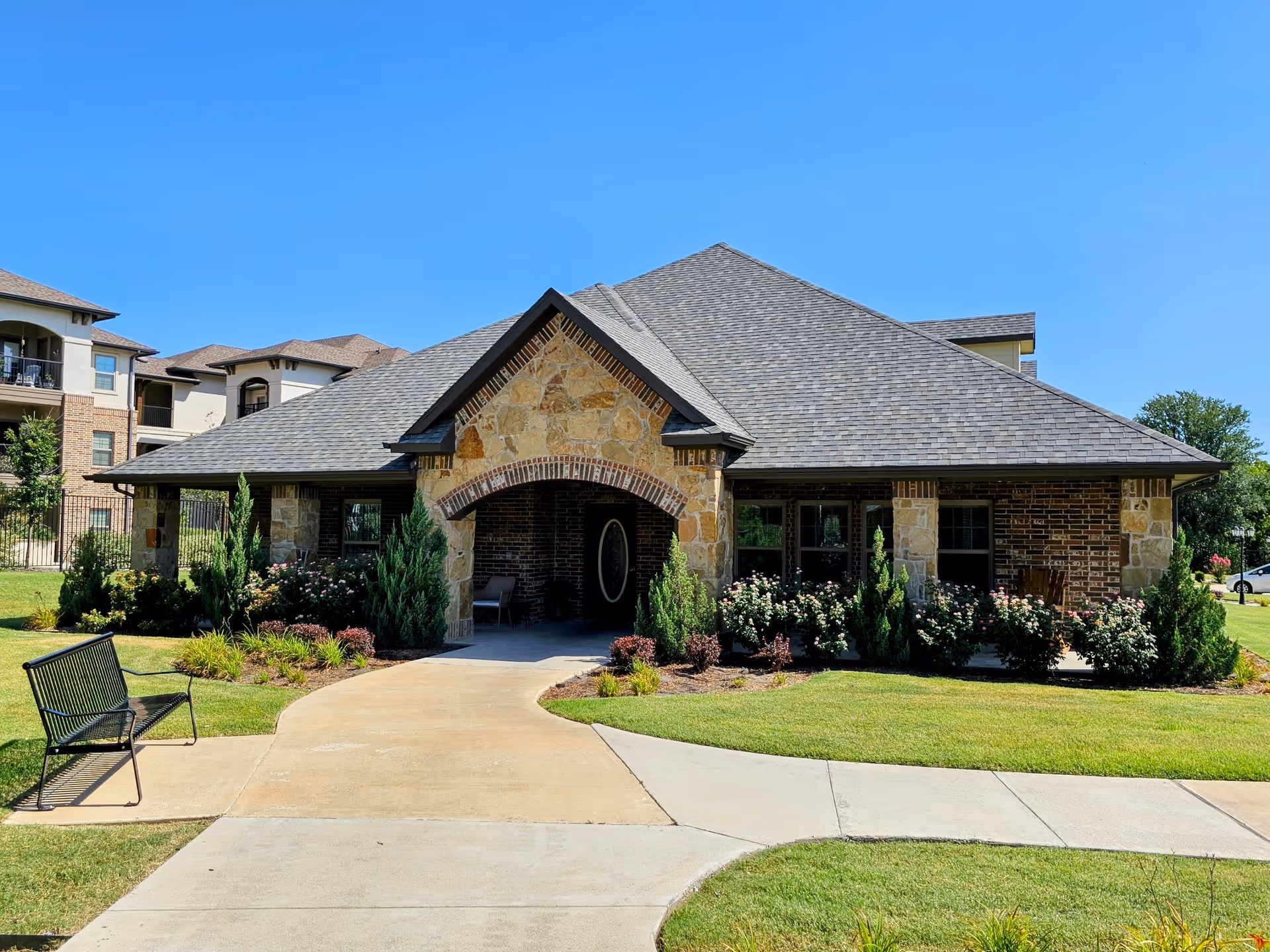 Front exterior view of a single-story assisted living facility building with a stone and brick facade, a covered entrance with an arched roof, surrounded by well-maintained landscaping including bushes and flowers, and a bench on the sidewalk leading to the entrance under a clear blue sky.
