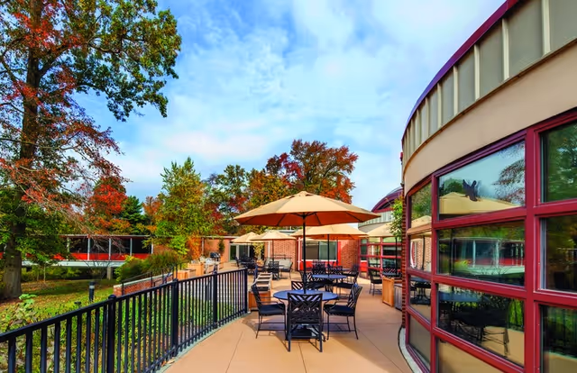 Outdoor patio area at Meadow Lakes with several tables and chairs under large beige umbrellas. The patio is adjacent to a building with large windows framed in red. Surrounding the patio are trees with autumn foliage and a black metal railing along the edge.