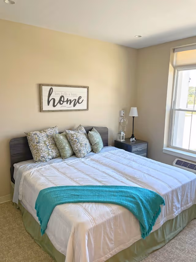 A neatly made bed with white bedding, a teal throw blanket at the foot, and five decorative pillows in floral and green patterns. Above the bed is a framed sign that reads 'love builds a happy home.' To the right of the bed is a nightstand with a lamp, a small lantern, and a decorative item. A large window with a view outside is on the right wall, letting in natural light.