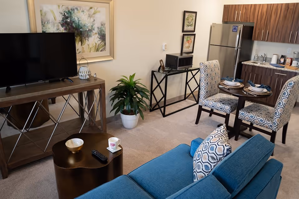 Open-plan living area with a teal sofa, TV on a console, a small dining table and kitchenette.