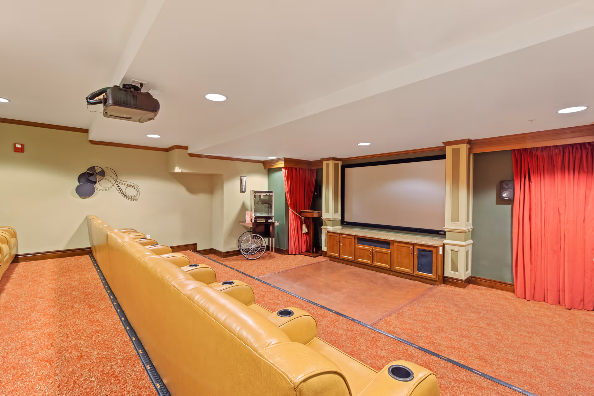 A small theater room with two rows of tan leather seats facing a large projection screen. The room has red carpet, red curtains on either side of the screen, a popcorn machine in the corner, and a ceiling-mounted projector.