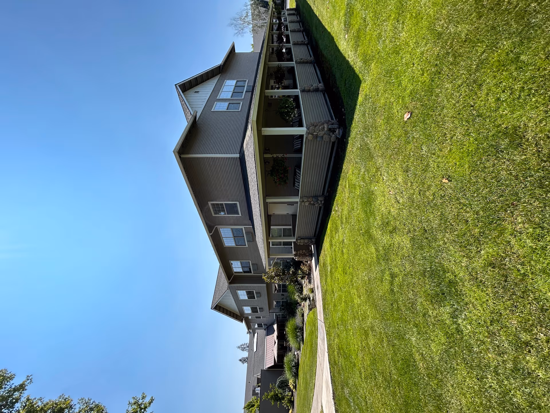 Exterior view of a two-story senior living facility building with a covered porch, surrounded by a well-maintained green lawn and landscaping under a clear blue sky.