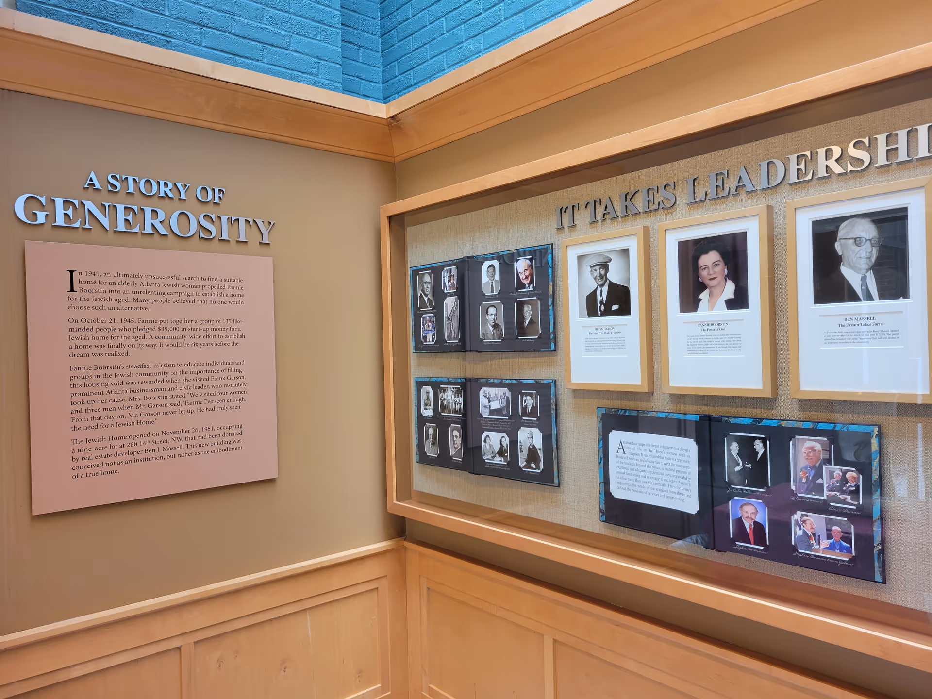 A wall display in a facility featuring a story titled 'A Story of Generosity' with text about Fannie Boorstin and the founding of a home for elderly Atlanta Jewish women. The display also includes framed photographs and text under the heading 'It Takes Leadership.' The wall is beige with wooden paneling and a blue brick upper section.