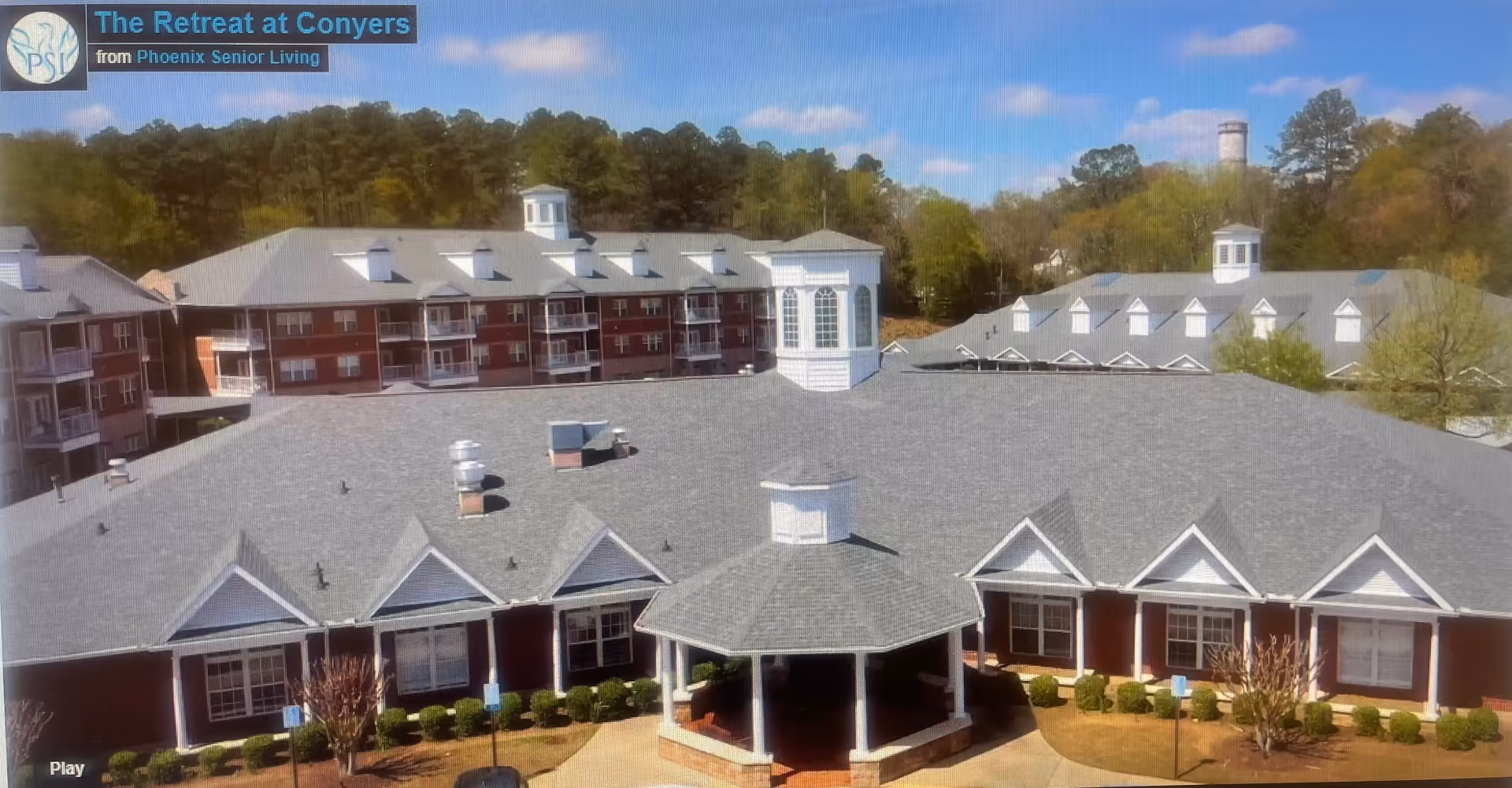 Aerial view of The Retreat at Conyers senior living facility showing multiple connected buildings with gray roofs, white trim, and red brick walls. The foreground features a covered gazebo structure surrounded by landscaped bushes and small trees. The background includes a wooded area under a blue sky with scattered clouds.