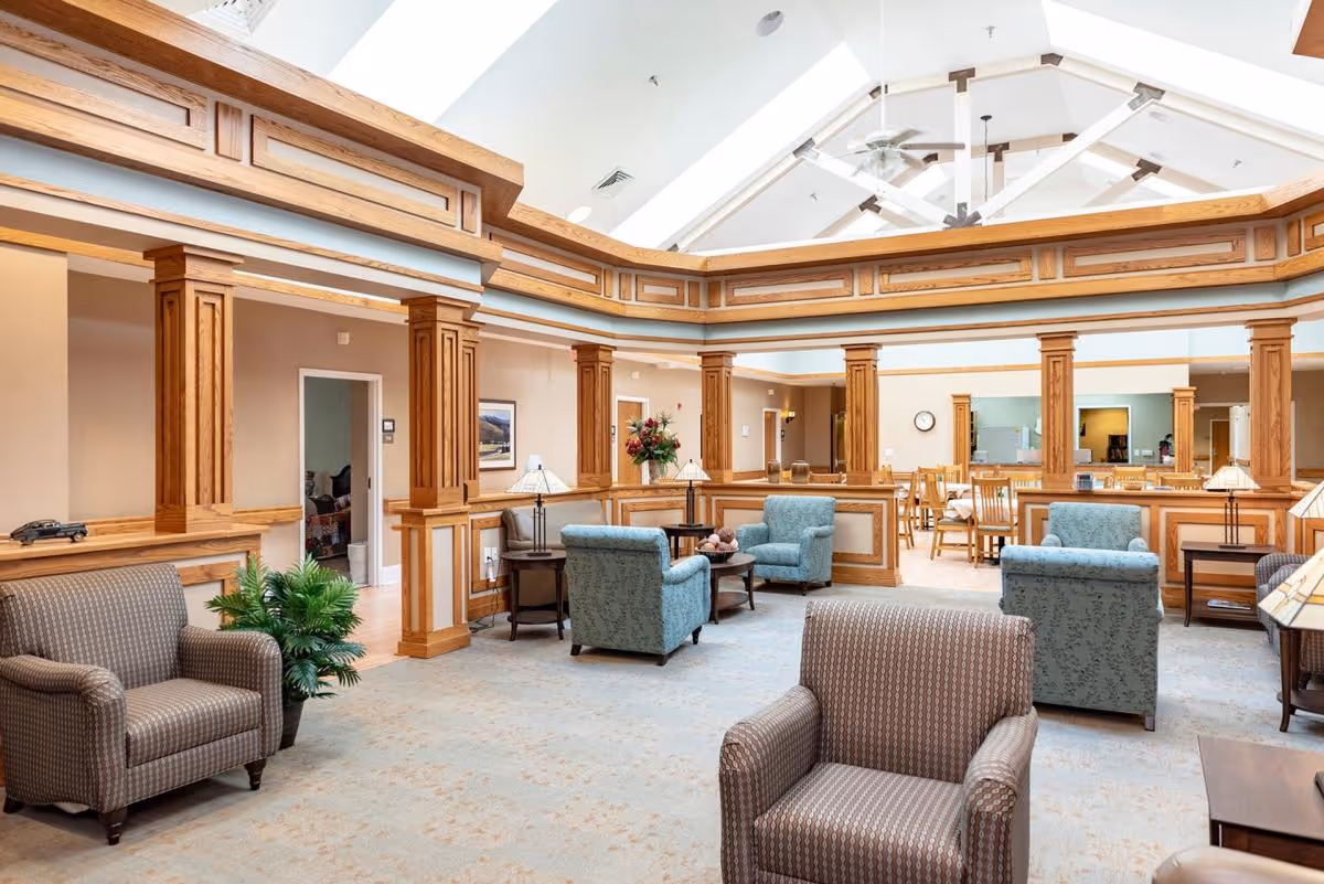 A spacious senior living facility common area with comfortable armchairs and small tables arranged for conversation. The room features wooden columns and trim, a high vaulted ceiling with ceiling fans, and a dining area with tables and chairs visible in the background.