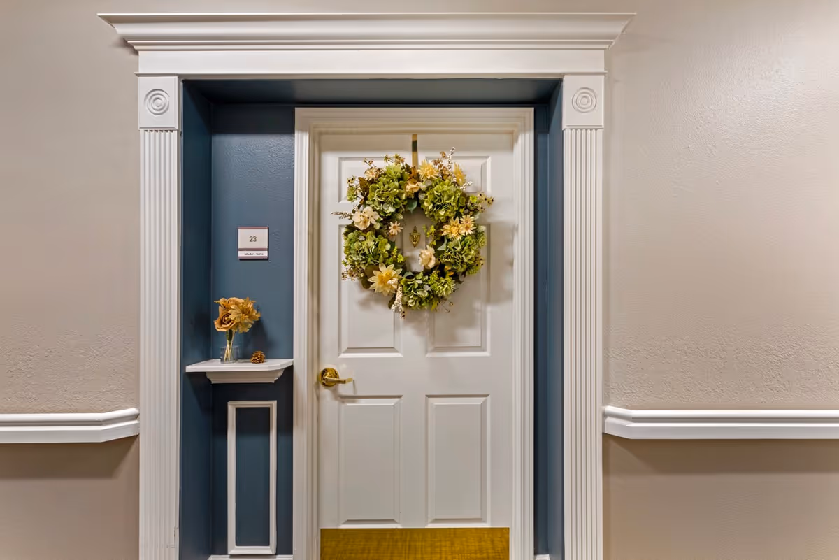 White apartment door in a senior living hallway decorated with a floral wreath and a small shelf with flowers and a room number plaque.