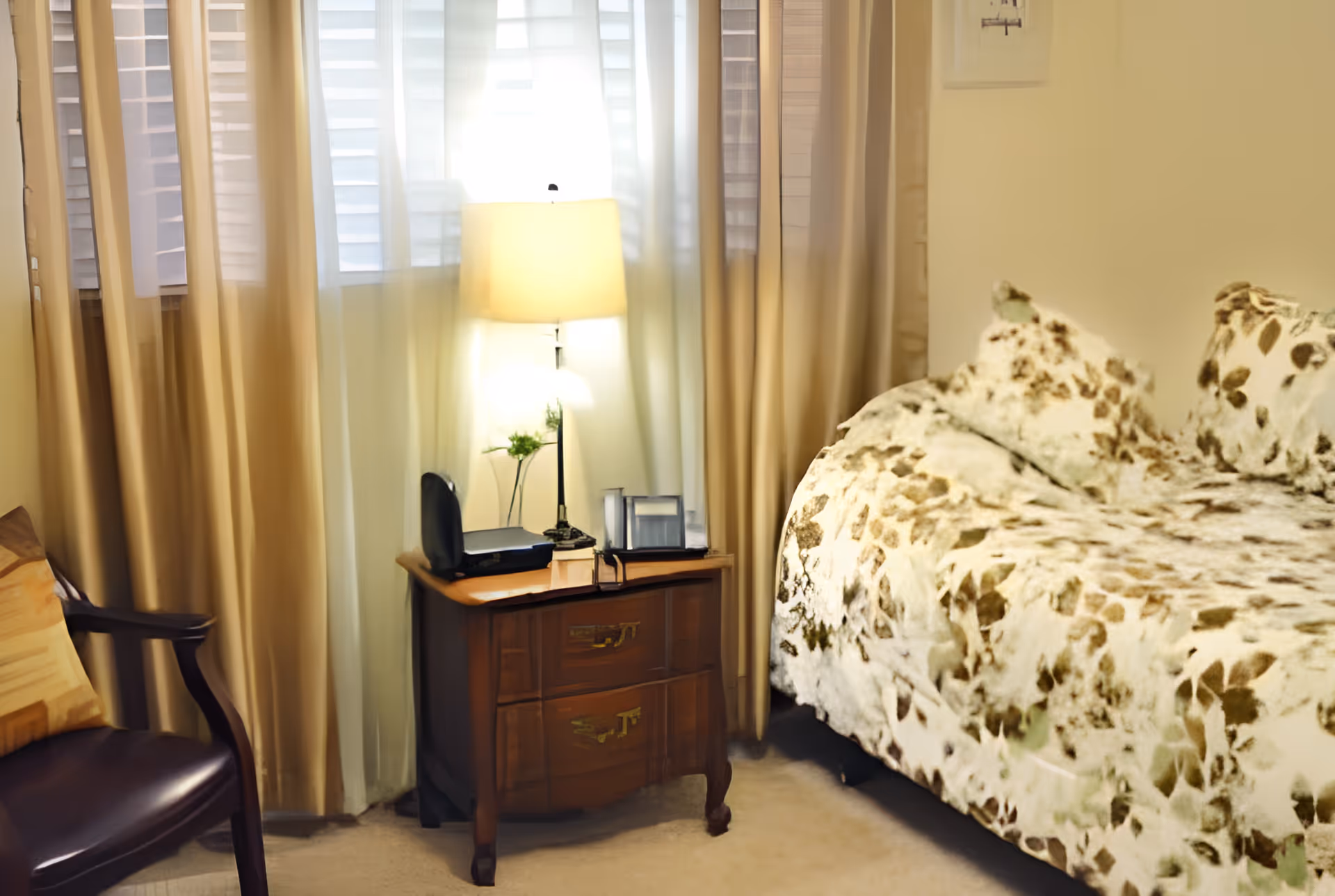 A cozy bedroom with a floral-patterned bedspread and matching pillows on the right side. Next to the bed is a wooden nightstand with two drawers, holding a table lamp, a small plant, and some items. Beige curtains cover the windows behind the nightstand, and a dark wooden chair with a cushion is partially visible on the left.