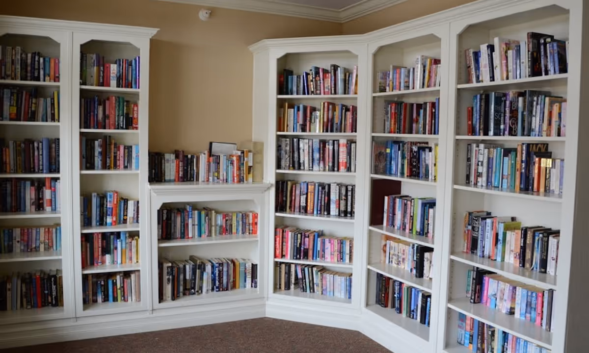 A corner of a room with white built-in bookshelves filled with a variety of books. The shelves are neatly organized and the room has beige walls and a brown carpeted floor.