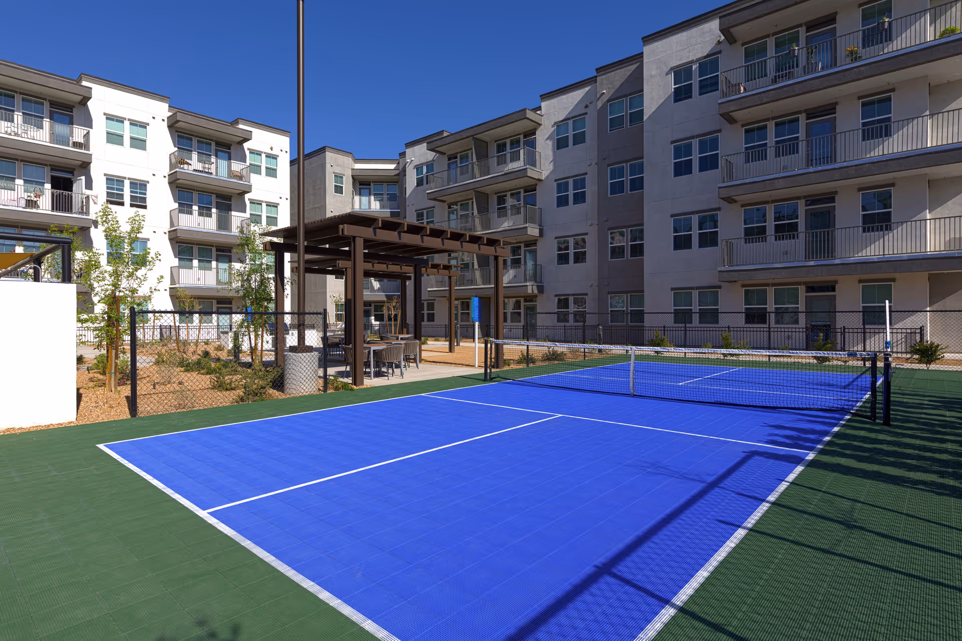 Outdoor view of a senior living facility featuring a blue and green pickleball court with a net, surrounded by a black chain-link fence. In the background, there are multi-story residential buildings with balconies and windows. A shaded seating area with chairs and tables is also visible near the court under a wooden pergola. The sky is clear and blue.